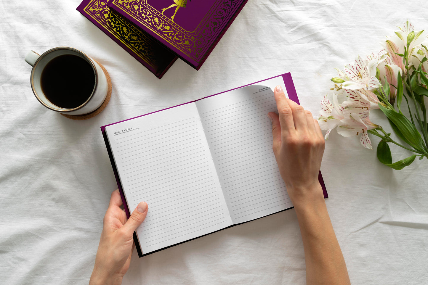 Person holding an open notebook with a cup of coffee and flowers on a white surface