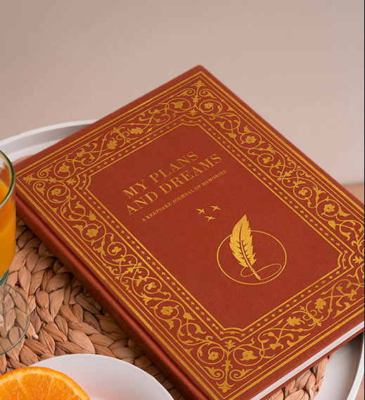 Vintage book titled 'My Plans and Dreams' with a glass of orange juice and orange slices on a woven tray.